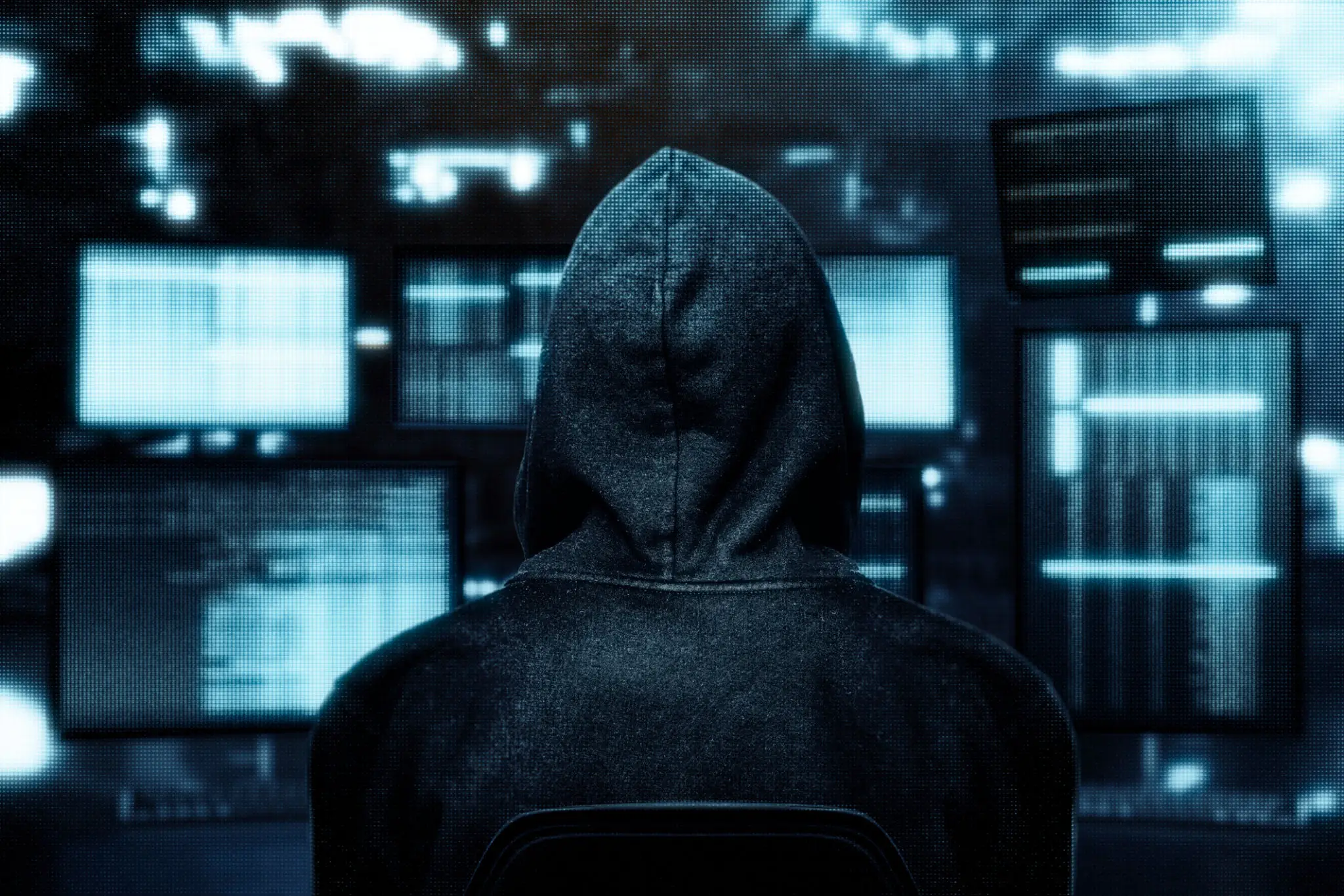 Hacker in hooded jacket at computer screens.